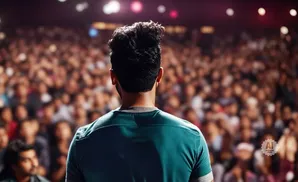 A man stands with his back to the camera, facing a large, blurred audience in a darkened venue.