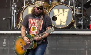 Musician playing an acoustic guitar on stage with drums in the background.