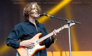 A masked guitarist plays an orange electric guitar on stage under yellow lights.