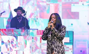 A female singer in a leopard-print coat holds a microphone on stage, with a DJ in a hat behind her.