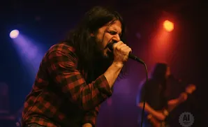 A man with long dark hair sings passionately into a microphone on stage, wearing a red and black plaid shirt.
