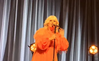 Singer with blonde hair in an orange dress and fur stole sings into a microphone on stage with white curtains and stage lights.