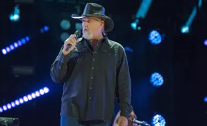 Man in a cowboy hat and black shirt sings into a microphone on a stage with blue lights.