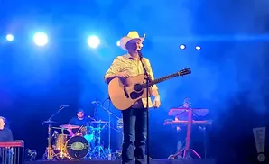 A man in a cowboy hat plays guitar on stage with a band, illuminated by blue stage lights.