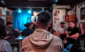 A man in a hoodie watches a performance on stage in a dimly lit venue.