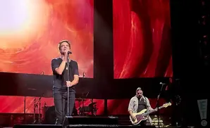 A man sings into a microphone on stage, while another plays bass guitar. Red and orange screens are behind them.