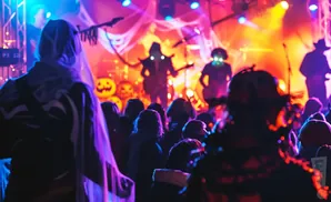 A crowd of people in costumes watch a band on a stage illuminated by colorful Halloween-themed lights and decorations.