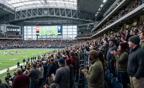 A football game is underway in a packed, modern stadium with a large screen showing the action.