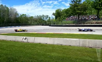 IndyCars race on a sunny track with spectators in the background.