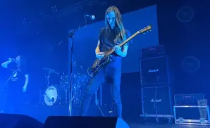 A rock band performs on a stage bathed in blue light, with a guitarist center stage.