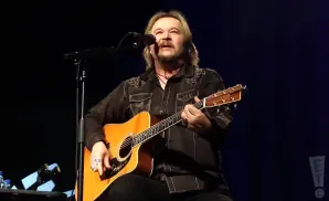 A man plays an acoustic guitar and sings into a microphone on a stage.