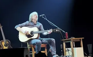 A musician with blonde hair plays an acoustic guitar while sitting on a stool on stage.