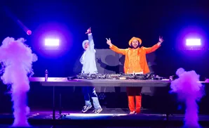 Two DJs in bright outfits enthusiastically perform on stage with purple smoke and vibrant blue lighting.