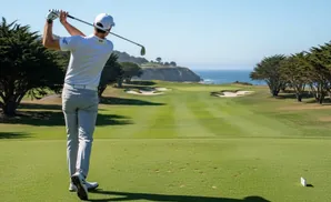 Golfer swings club on a scenic golf course with ocean views.