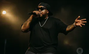 Rapper performs on stage, wearing a black t-shirt, black baseball cap, sunglasses, and jewelry, holding a microphone.