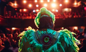 A person in a bright green feathered costume stands facing away from the camera, with a crowd and stage lights in the background.