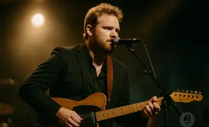 A man with red hair and a beard plays a guitar and sings into a microphone.