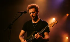 A man with curly red hair plays an electric guitar on stage, illuminated by warm stage lights.