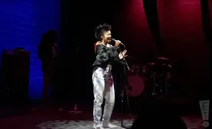 Singer performing on stage with a blue brick wall in the background and red stage lights.