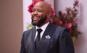 A bald, bearded Black man in a sparkly blue suit smiles widely.
