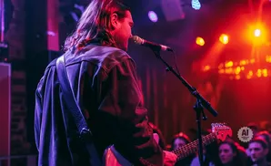 A man with long hair plays guitar and sings into a microphone on a dimly lit stage with red and purple lights.
