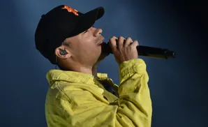 A man in a yellow jacket and black cap sings into a microphone with an earbud in his ear.