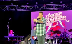 A male singer in a plaid shirt performs on stage with a band behind him.