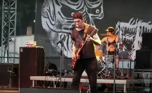 A band performs on stage, with a guitarist singing into a microphone, and a drummer behind them.