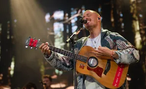 A man sings and plays a guitar on stage, wearing a patterned jacket and necklaces.