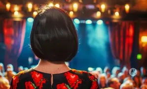 Back of a woman's head with short dark hair and a black dress with red floral print, facing a stage with lights and a blurred audience.
