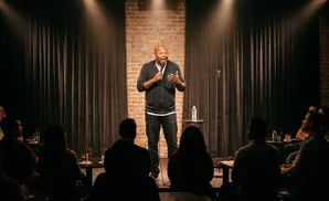 A Black comedian tells a joke on stage to a seated audience in a dimly lit club.
