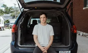 Man in white t-shirt sits in the open trunk of a dark minivan, looking at the camera.