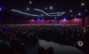 An audience watches a presentation on a large screen in a dimly lit auditorium with purple curtains.