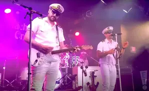 Musicians in white captain's uniforms perform on stage under purple lights, one playing guitar, the other a tambourine.