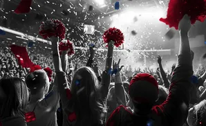 Crowd at a concert or game throwing red and blue confetti, with some holding red pom-poms.