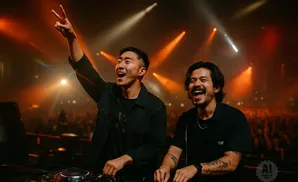 Two DJs laugh on stage at a concert with bright orange spotlights and a cheering crowd.