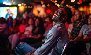 A man laughs heartily in a dimly lit room, with an audience in the background.