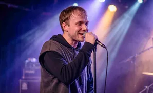 A male singer sweats as he performs into a microphone on a dimly lit stage.