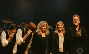 A musical group of five people on stage with microphones and drums in the background.