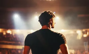 Man with curly hair on stage, facing away from camera, bathed in spotlight.