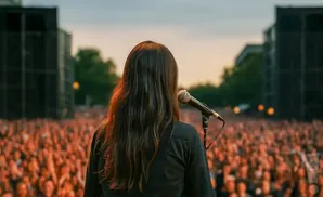 A person with long, dark hair speaks into a microphone on stage to a large, cheering crowd.