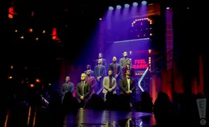 A group of men in suits stand on a stage with dramatic lighting.