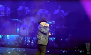 A man in a cowboy hat sings into a microphone on a stage with a band behind him.
