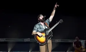 A man in a plaid shirt and baseball cap plays guitar and sings on stage, raising his hand.