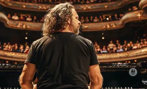A man with long, curly hair plays a grand piano on a stage in front of a blurred audience in a theater.