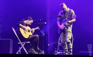 Two guitarists perform on a dimly lit stage, bathed in purple light.