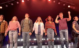 A group of seven young people in casual clothes stand onstage, holding hands and looking up.