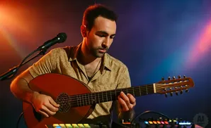 A musician plays a guitar with a microphone and music equipment in front of a colorful background.