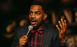 Man in a suit and bowtie speaking into a microphone, gesturing with his hand.