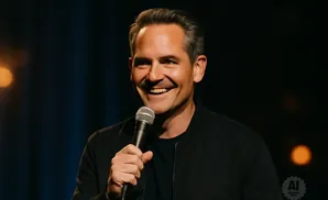 A man in a black jacket laughs while holding a microphone.